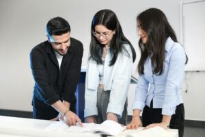 Trois jeunes adultes discutent et pointent du doigt des documents ouverts dans un bureau moderne, favorisant le travail d'équipe.