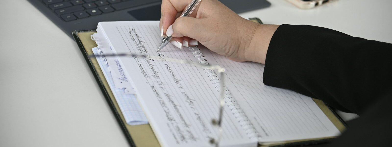 A person writing on a notebook with a laptop in the background